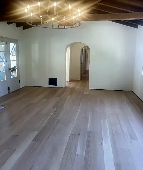 A white room with dark beams, an iron chandelier and an arched doorway, and a pale natural wood floor