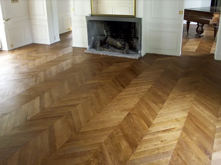 A chevron wood floor in warm brown in front of a white fireplace, the points lining up in rows across the room