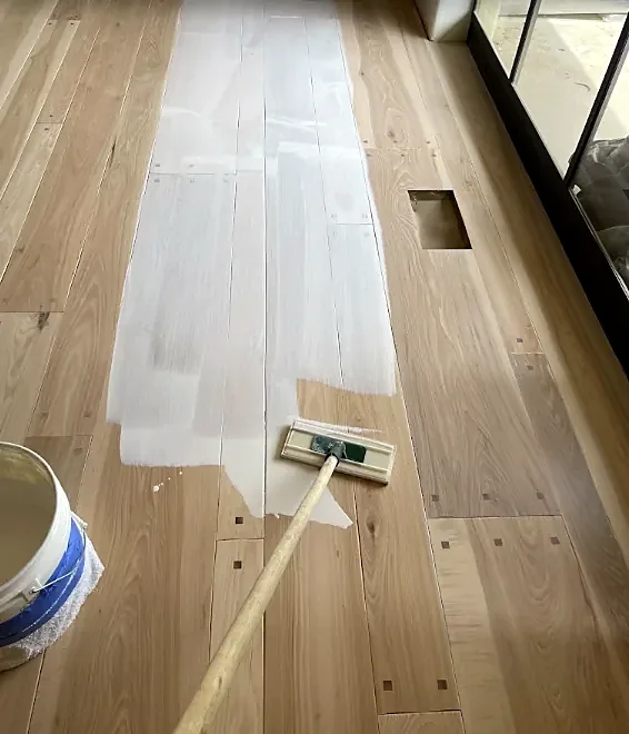 A wide applicator on a pole spreads a milky first coat down a run of pale new boards, with a bucket beside it