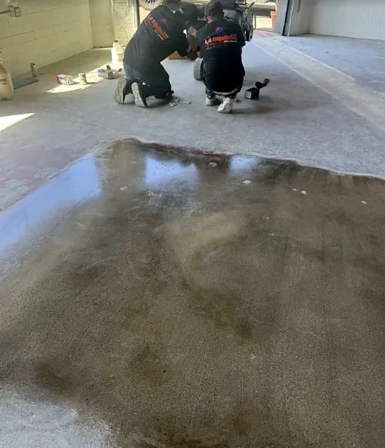 Two crew members in La Cumbre shirts kneel at the edge of a wet coating spread on a concrete floor
