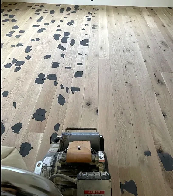 A pale new plank floor dotted with dark filler patches, with a drum sander in the foreground