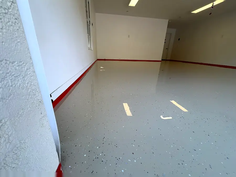 Una capa de epóxico fresca en un garaje con hojuelas oscuras esparcidas, cinta roja junto a la pared y papel azul en la orilla