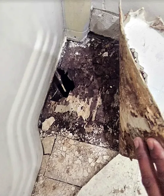 A hand peels back old flooring in a corner to show dark, crumbling material underneath