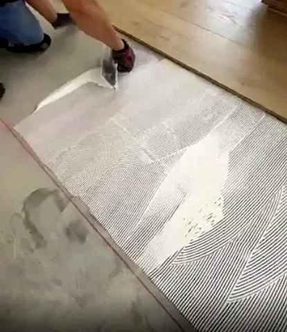 A gloved hand combs white adhesive into ridges with a notched trowel, next to the last row of planks