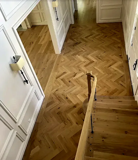 A herringbone oak floor runs down a paneled hall and meets the top step of a stair