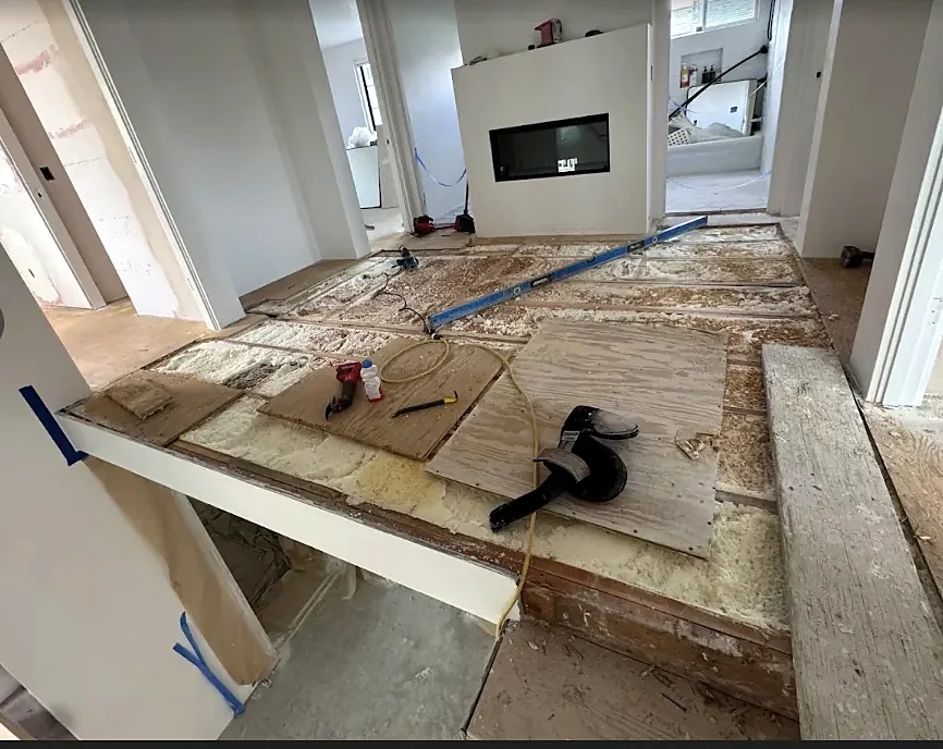 A floor opened down to the joists, with spray foam between them, plywood pieces, tools and a long level laid across