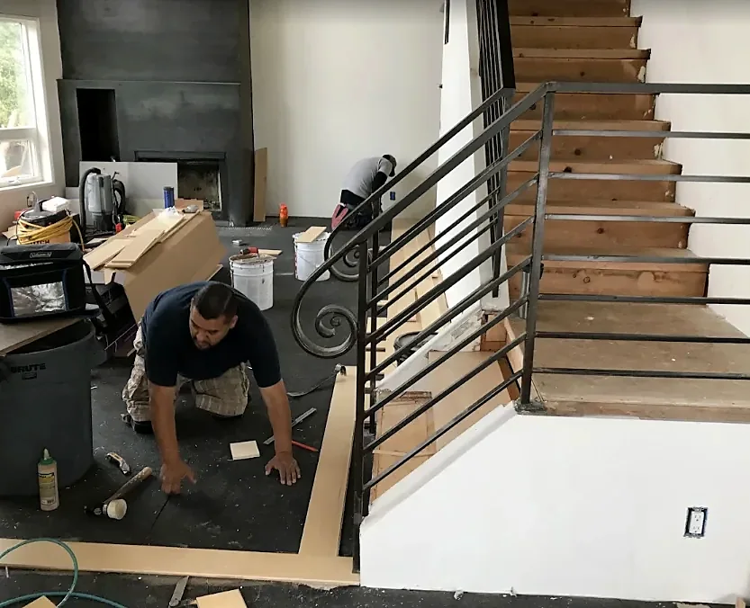 Two crew members fit border boards on the floor at the foot of a stair with an iron railing, tools and adhesive around them