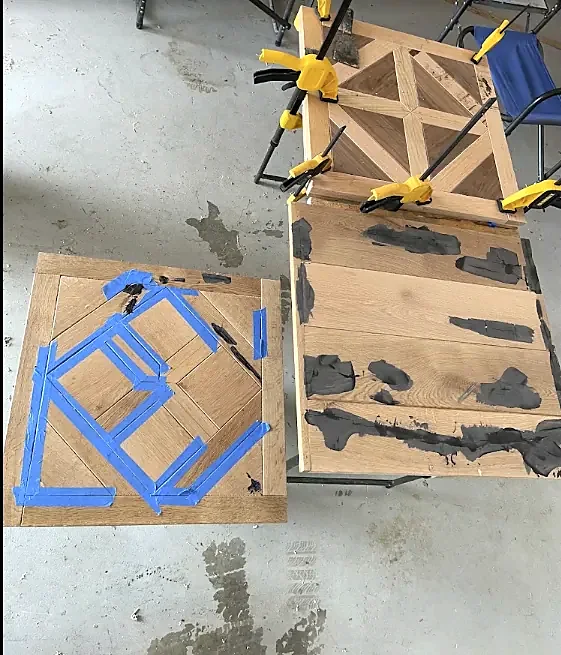 Parquet panels during glue-up on a garage floor: one held with blue tape, one in clamps