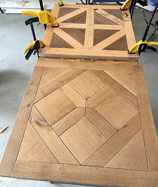 Two square parquet panels on a workbench: one finished in oak, one clamped in yellow clamps with darker wood triangles