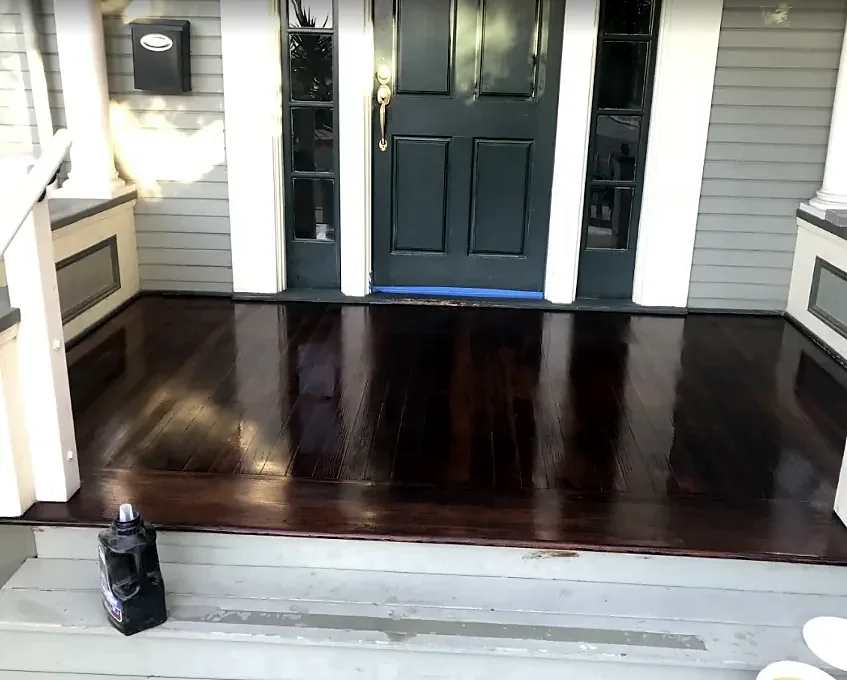 A front porch floor stained very dark and still wet with finish, with blue tape along the door sill