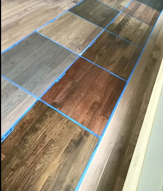Ten stain samples in different browns, grays and a red, each boxed in blue tape on a sanded floor
