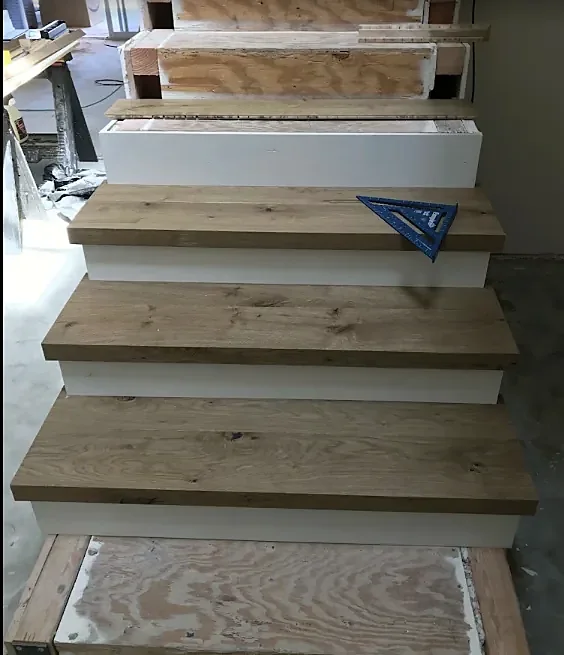 A stair halfway through: thick oak treads on the lower steps, bare plywood above, a speed square resting on one tread