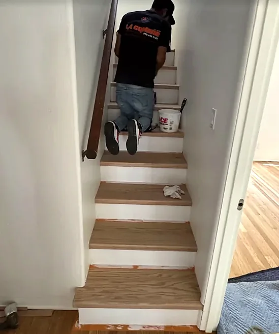 A crew member in a La Cumbre shirt kneels on a narrow stair, fitting oak treads over white risers