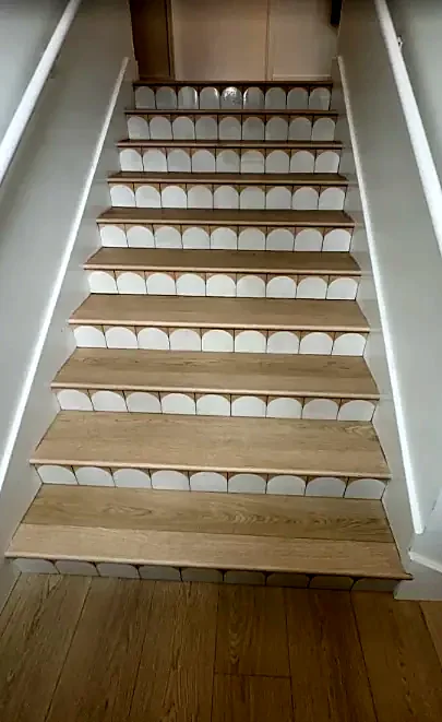 A straight stair seen from the bottom, with light oak treads and scalloped white tile risers