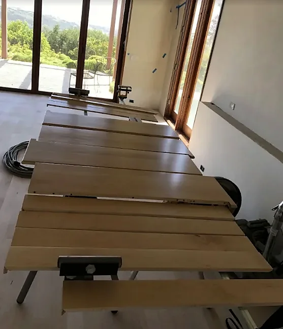 Wide boards laid out on sawhorses in a sunlit room beside glass doors