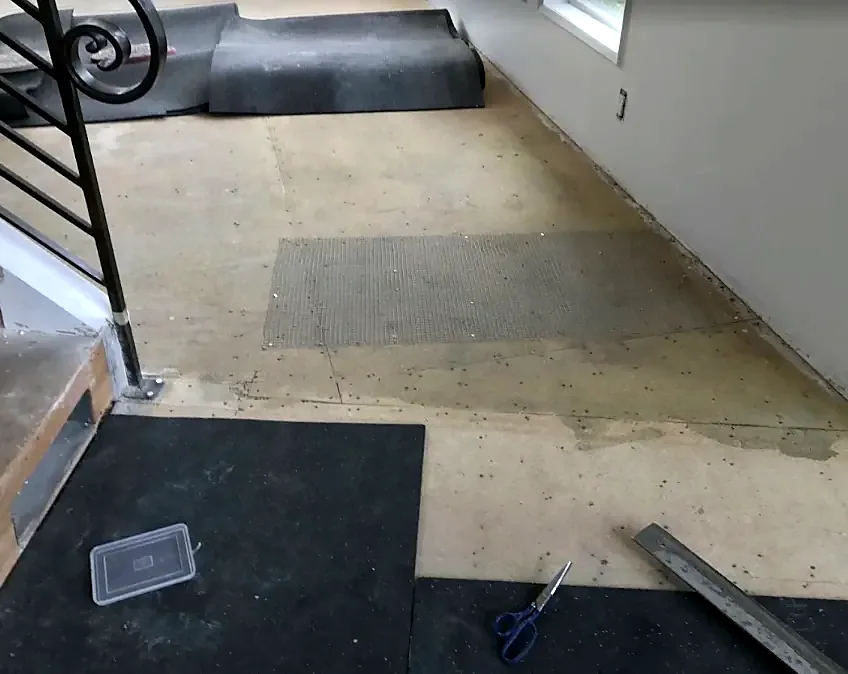 A bare subfloor beside an iron stair rail, with a patch of metal mesh and a long straightedge on the ground