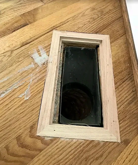 A rectangular vent opening in an oak floor with a new unfinished wood frame fitted around it