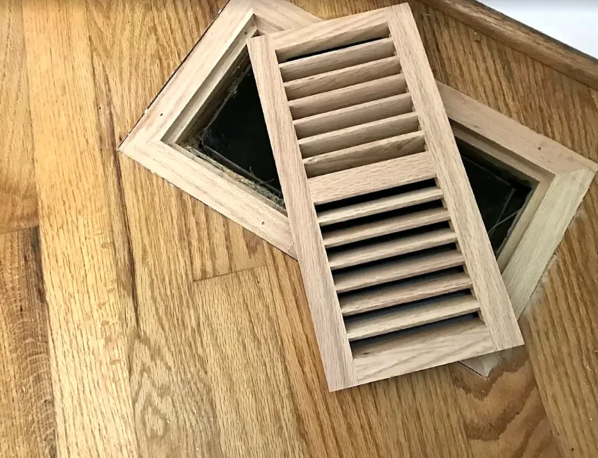 An unfinished wood vent frame and louvered insert resting on an oak floor over the vent opening